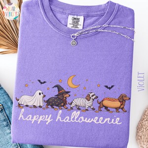 May include: A violet crewneck t-shirt featuring a graphic of four dachshunds in Halloween costumes, including a ghost, a witch, a mummy, and a hot dog, with a crescent moon, stars, bats, and the text "happy halloweenie" printed in white.