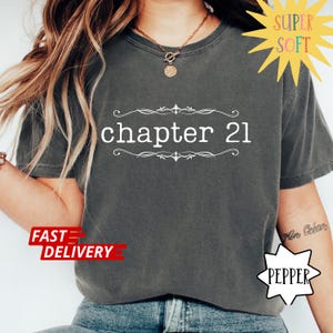 May include: Dark gray t-shirt with the text "chapter 21" in white, framed by decorative flourishes. The shirt has a crew neck and short sleeves. The words "Super Soft" and "Fast Delivery" are also visible.