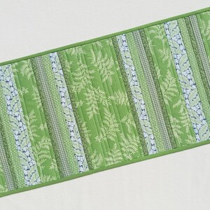 May include: Green and white quilted table runner with a fern leaf pattern. The runner has vertical stripes of varying patterns and shades of green, with a solid green border. The overall design is rectangular.