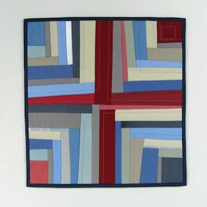 May include: A square quilt with a geometric pattern of red, blue, and gray fabric squares. The quilt is framed with a dark blue border.
