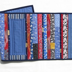 May include: A blue and red striped table runner with a patchwork design. The runner is made of various fabrics, including blue, red, white, and gold. It has a blue fabric panel in the center.