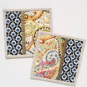 Puede incluir: Dos posavasos cuadrados de tela. Uno presenta un fondo negro con un estampado floral azul y blanco, una franja beige y un diseño de cachemira. El otro posavasos tiene un diseño similar. Ambos están bordeados con un estampado floral.