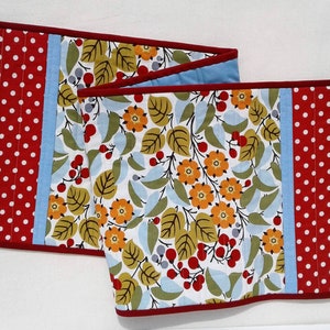 May include: Two red and white polka dot fabric table runners with a blue stripe and a red border. The runners are decorated with a floral pattern of green leaves, orange and yellow flowers, and red berries.