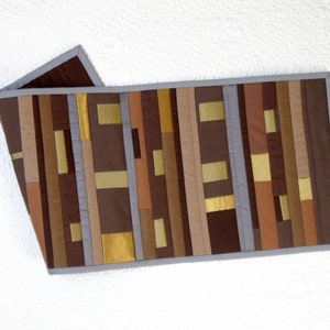 May include: A rectangular table runner with a patchwork design. The runner features vertical stripes in shades of brown, tan, and gold, with gray accents. The edges are finished with a gray border. The runner is made of fabric.