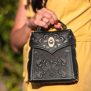 Könnte beinhalten: Ein schwarzer Lederrucksack mit aufwendigen geprägten Schnörkeln und einem messingfarbenen Verschluss. Die Tasche hat einen oberen Griff. Der Rucksack wird von einer Person in einem gelben Kleid gehalten. Das Design ist detailliert und verziert.