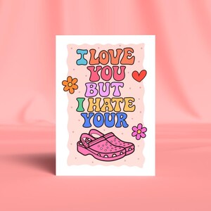 May include: A greeting card with the text "I LOVE YOU BUT I HATE YOUR" in colourful bubble letters. A pink cartoon clog is at the bottom, with a red heart and flowers on the sides. The background is light pink.