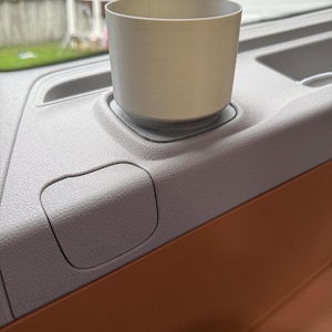 Op de afbeelding: Een lichtgrijze bekerhouder ontworpen voor het interieur van een voertuig. De bekerhouder is cilindrisch en staat op een basis, geplaatst in het dashboard van een voertuig. Het dashboard is lichtgrijs, met een bruin accent. De achtergrond toont een huis en een garage.