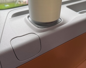 Op de afbeelding: Een lichtgrijze bekerhouder ontworpen voor het interieur van een voertuig. De bekerhouder is cilindrisch en staat op een basis, geplaatst in het dashboard van een voertuig. Het dashboard is lichtgrijs, met een bruin accent. De achtergrond toont een huis en een garage.