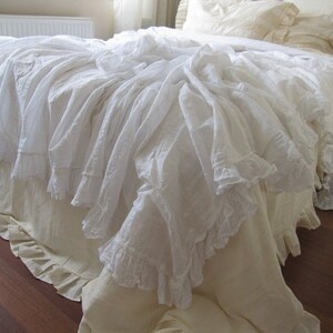 May include: White ruffled bed skirt with a white ruffled bedspread. The bedspread is draped over the bed skirt and has a lot of ruffles.