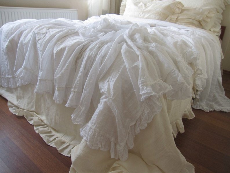 May include: White ruffled bed skirt with a white ruffled bedspread. The bedspread is draped over the bed skirt and has a lot of ruffles.