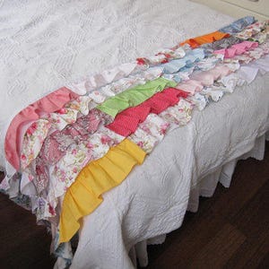 May include: A white bedspread with a colorful ruffled trim. The trim is made of different fabrics in shades of pink, red, orange, yellow, green, blue, and purple. The trim is arranged in a cascading pattern, with each ruffle overlapping the one below it.