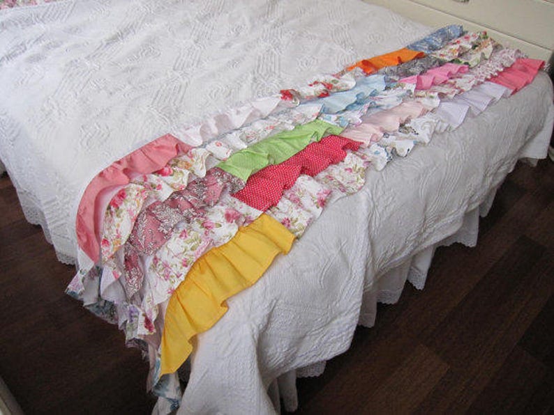 May include: A white bedspread with a colorful ruffled trim. The trim is made of different fabrics in shades of pink, red, orange, yellow, green, blue, and purple. The trim is arranged in a cascading pattern, with each ruffle overlapping the one below it.