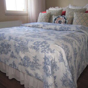 May include: A white bed with a blue and white toile patterned duvet cover and a white lace bed skirt. The bed is made with several pillows, including a floral patterned pillow.