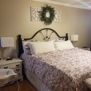 May include: A bedroom scene featuring a black metal bed frame with a floral patterned comforter and white pillows. A decorative wreath hangs above the bed. White nightstands with lamps flank the bed, and a baby bassinet is visible.
