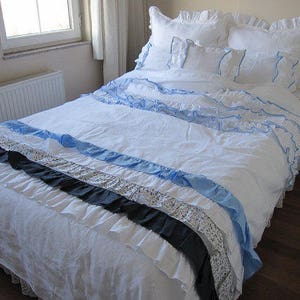 May include: A white bed with a white and blue ruffled bedspread. The bedspread has a black and white patterned ruffle at the bottom. The bed is made with white pillows and a white and blue patterned pillow.