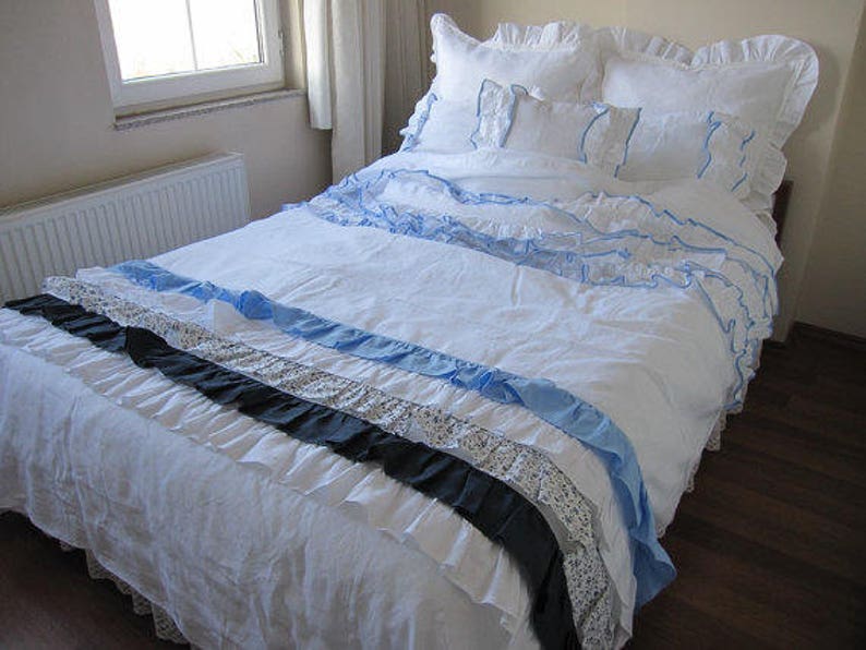 May include: A white bed with a white and blue ruffled bedspread. The bedspread has a black and white patterned ruffle at the bottom. The bed is made with white pillows and a white and blue patterned pillow.