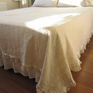 May include: A white bedspread with a ruffled edge and a crocheted lace trim. The bedspread is made of a soft, textured fabric and has a slightly distressed look.