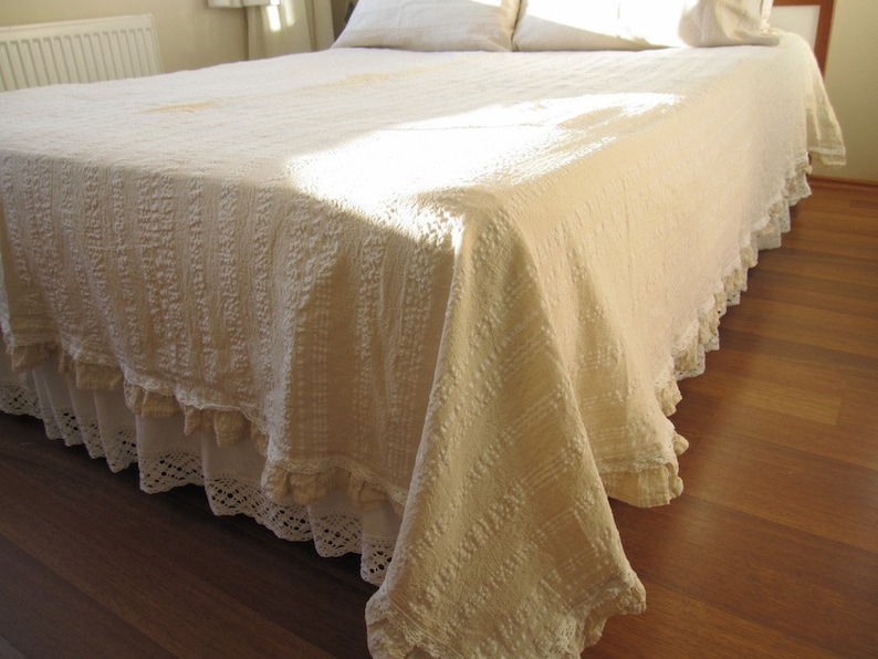 May include: A white bedspread with a ruffled edge and a crocheted lace trim. The bedspread is made of a soft, textured fabric and has a slightly distressed look.