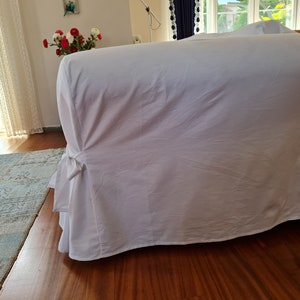 May include: White sofa slipcover with a gathered skirt and a tied bow detail. The slipcover is made of a soft, smooth fabric.