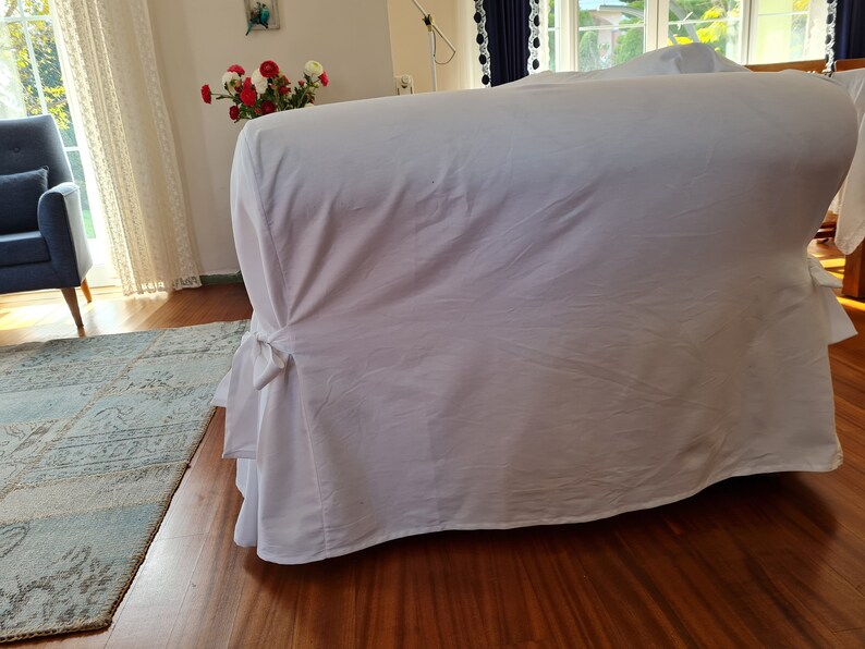 May include: White sofa slipcover with a gathered skirt and a tied bow detail. The slipcover is made of a soft, smooth fabric.