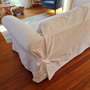 May include: A white sofa with a white slipcover and a bow tie detail at the bottom. The sofa is in a living room with hardwood floors.