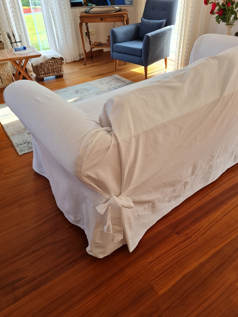 May include: A white sofa with a white slipcover and a bow tie detail at the bottom. The sofa is in a living room with hardwood floors.