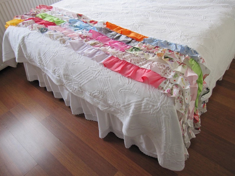May include: A white bedspread with a white ruffled bed skirt. The bedspread has a colorful patchwork of fabric strips with ruffles sewn along the edges.