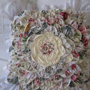 May include: A decorative pillow with a large, cream-colored flower in the center surrounded by smaller, multi-colored floral fabric rosettes.