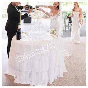 May include: A wedding reception scene with a tiered champagne glass display on a round table covered in a white ruffled cloth. A bride and groom are pouring champagne into the glasses. The table has the text "NURDANCEYIZ" on the front.