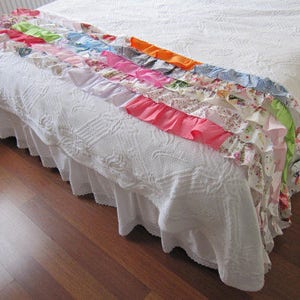 May include: A white bedspread with a ruffled trim and a colorful patchwork of fabric strips sewn along the top edge. The fabric strips are in various shades of pink, blue, green, orange, and yellow.