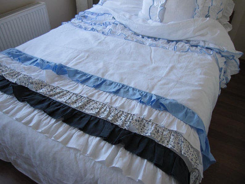 May include: A white bedspread with multiple layers of ruffles in blue, white, and black. The bedspread is made of a soft, linen-like fabric.
