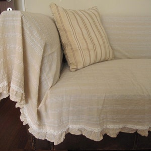 K&ouml;nnte beinhalten: Ein beigefarbenes Sofa mit einem wei&szlig;en und beige gestreiften Kissen und einer wei&szlig;en H&auml;keldecke mit einer Spitzenborte.