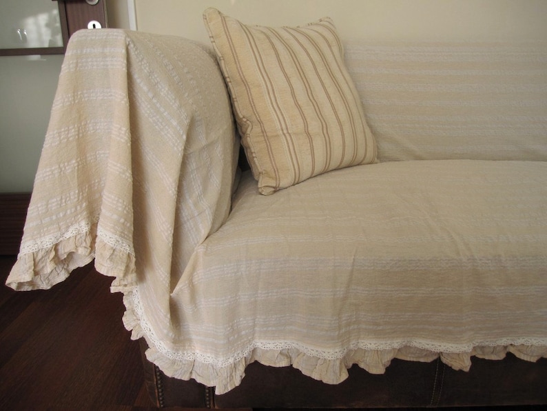 K&ouml;nnte beinhalten: Ein beigefarbenes Sofa mit einem wei&szlig;en und beige gestreiften Kissen und einer wei&szlig;en H&auml;keldecke mit einer Spitzenborte.