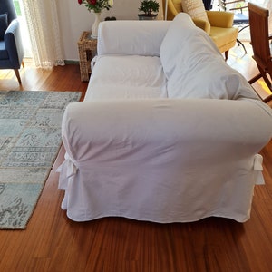 May include: A white sofa with a white slipcover and two pillows. The sofa is in a living room with a wooden floor and a rug.