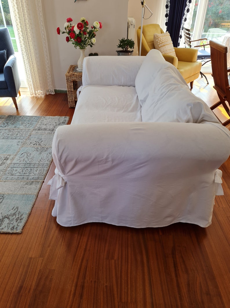 May include: A white sofa with a white slipcover and two pillows. The sofa is in a living room with a wooden floor and a rug.