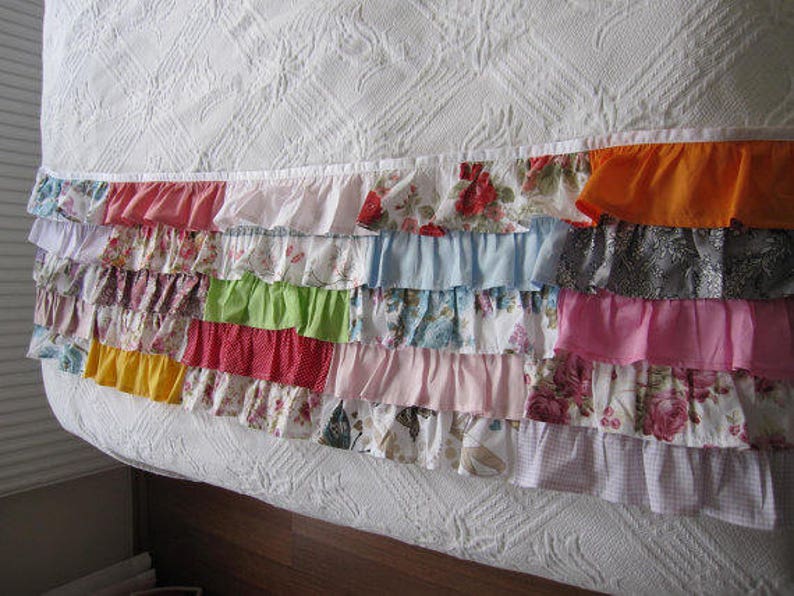 May include: A white bed with a colorful ruffled trim. The trim is made of various fabrics in different colors and patterns, including floral, plaid, and solid colors.