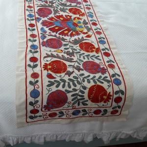 May include: A white table runner with a floral embroidery design. The embroidery features red, blue, green, and yellow flowers and leaves. The runner has a white lace trim.