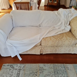 May include: A white sofa cover draped over a beige patterned sofa with a floral design. The sofa is in a living room setting with a wooden floor and a rug.