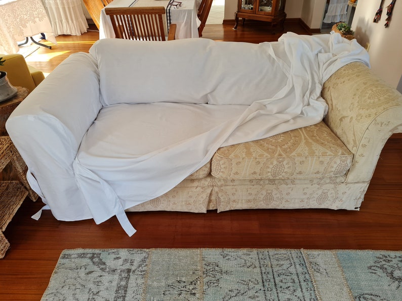 May include: A white sofa cover draped over a beige patterned sofa with a floral design. The sofa is in a living room setting with a wooden floor and a rug.