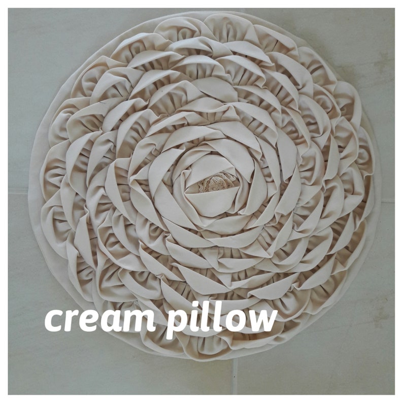 May include: Cream colored round pillow with a ruffled design. The pillow is made of fabric and has a soft, plush texture.