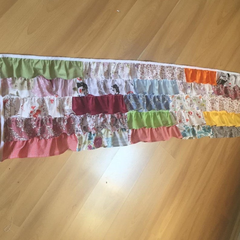 May include: A colorful patchwork valance with a ruffled bottom edge. The valance is made of various fabrics in shades of red, pink, green, blue, yellow, and orange. The fabric is sewn together in a random pattern, creating a unique and eye-catching design.
