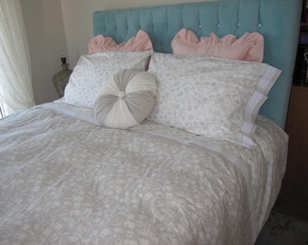 May include: A white and gray patterned bedspread with a white and gray round pillow on top of a bed with two white and gray patterned pillows and two pink ruffled pillows. The headboard is teal blue with a tufted design.