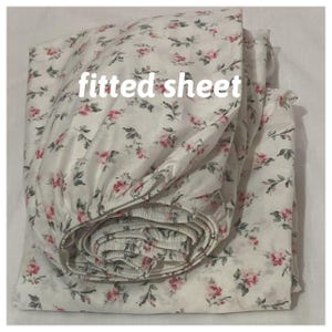 Pode incluir: Um lençol de baixo branco com um padrão floral de rosas cor-de-rosa e folhas verdes. O texto "fitted sheet" é impresso na parte superior do lençol em letras brancas. O lençol está dobrado e enrolado.