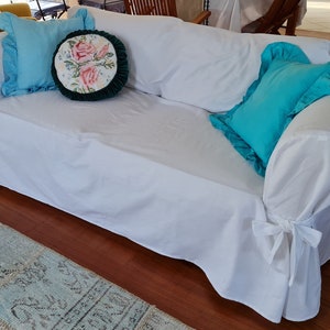 May include: White sofa slipcover with a ruffled edge and ties at the bottom. The slipcover is decorated with two pillows, one blue and one with a floral design.