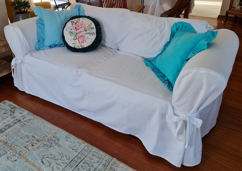 May include: White sofa slipcover with a ruffled edge and ties at the bottom. The slipcover is decorated with two pillows, one blue and one with a floral design.
