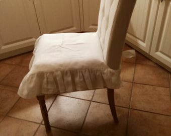 May include: A cream-colored upholstered dining chair with a high back and button tufting. The seat is covered with a white fabric, and the chair has a ruffled skirt. The legs are made of dark wood.