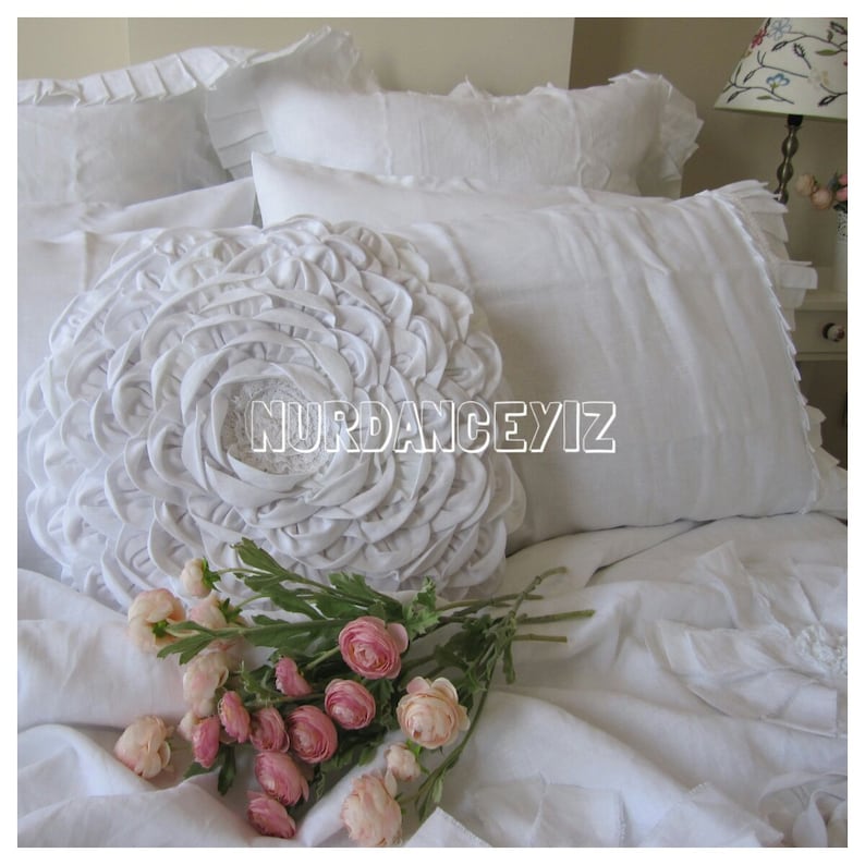 May include: White ruffled pillow with a large white flower-shaped pillow in the center. A bouquet of pink flowers is on the bed.
