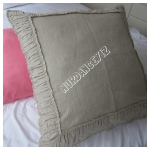 May include: A beige pillow with a ruffled edge. The pillow is made of a soft, textured fabric. The text "NURDANCEY/IZ" is embroidered on the pillow.
