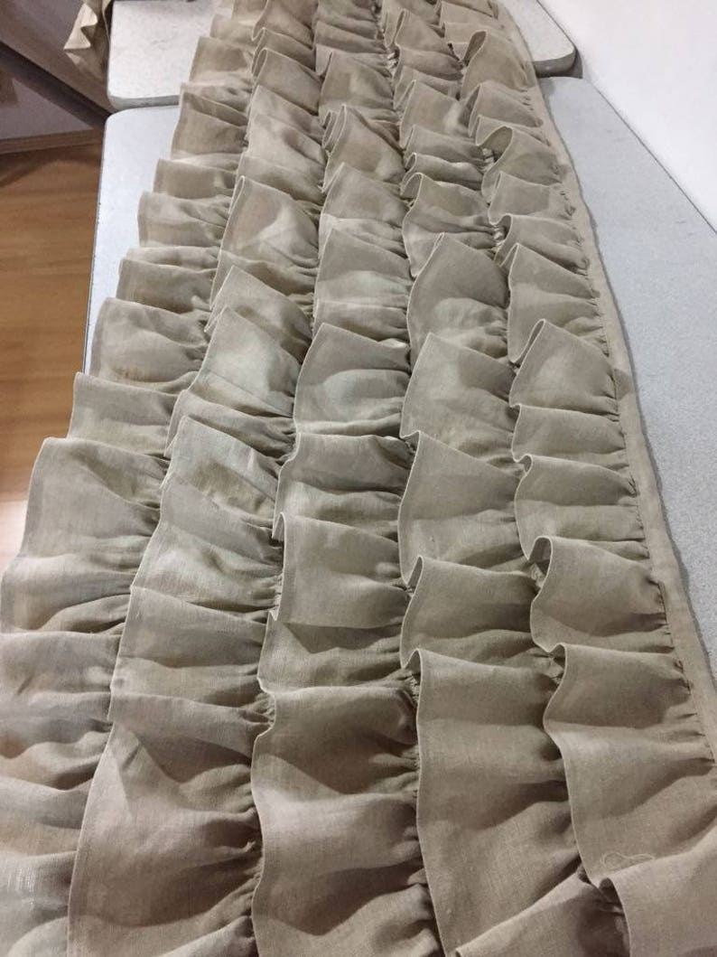 May include: A long, beige, ruffled fabric valance with multiple layers of ruffles. The fabric is a linen blend and has a slightly textured look.