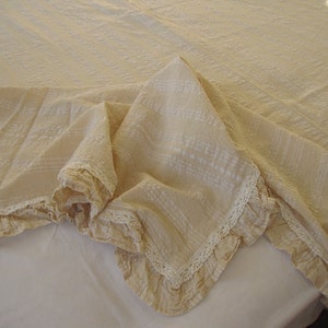 May include: A cream-colored throw blanket with a delicate lace trim. The blanket is made of a soft, textured fabric and has a subtle pattern.